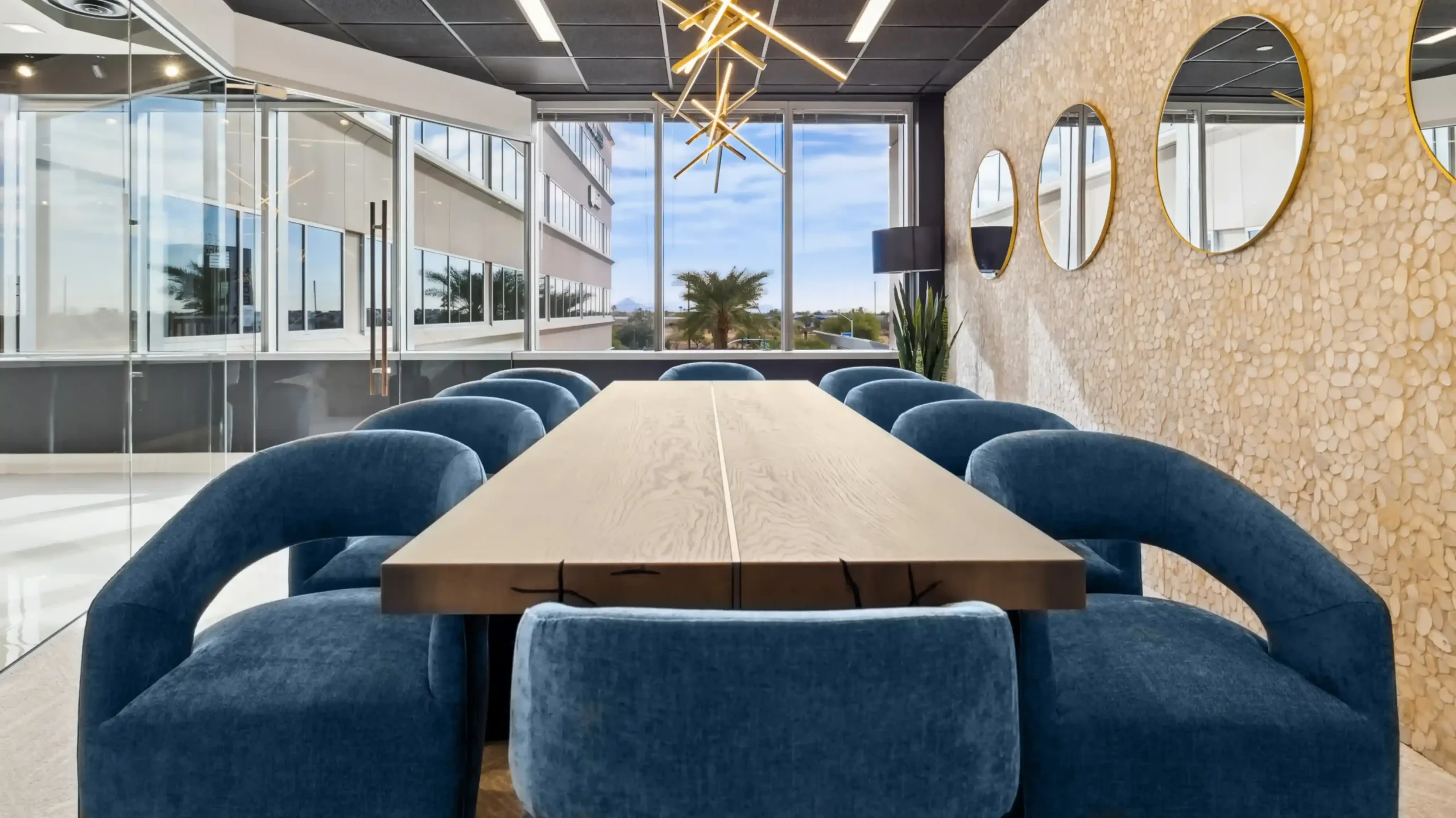 Long conference table with blue chairs beside floor-to-ceiling windows, with a round mirror on the wall