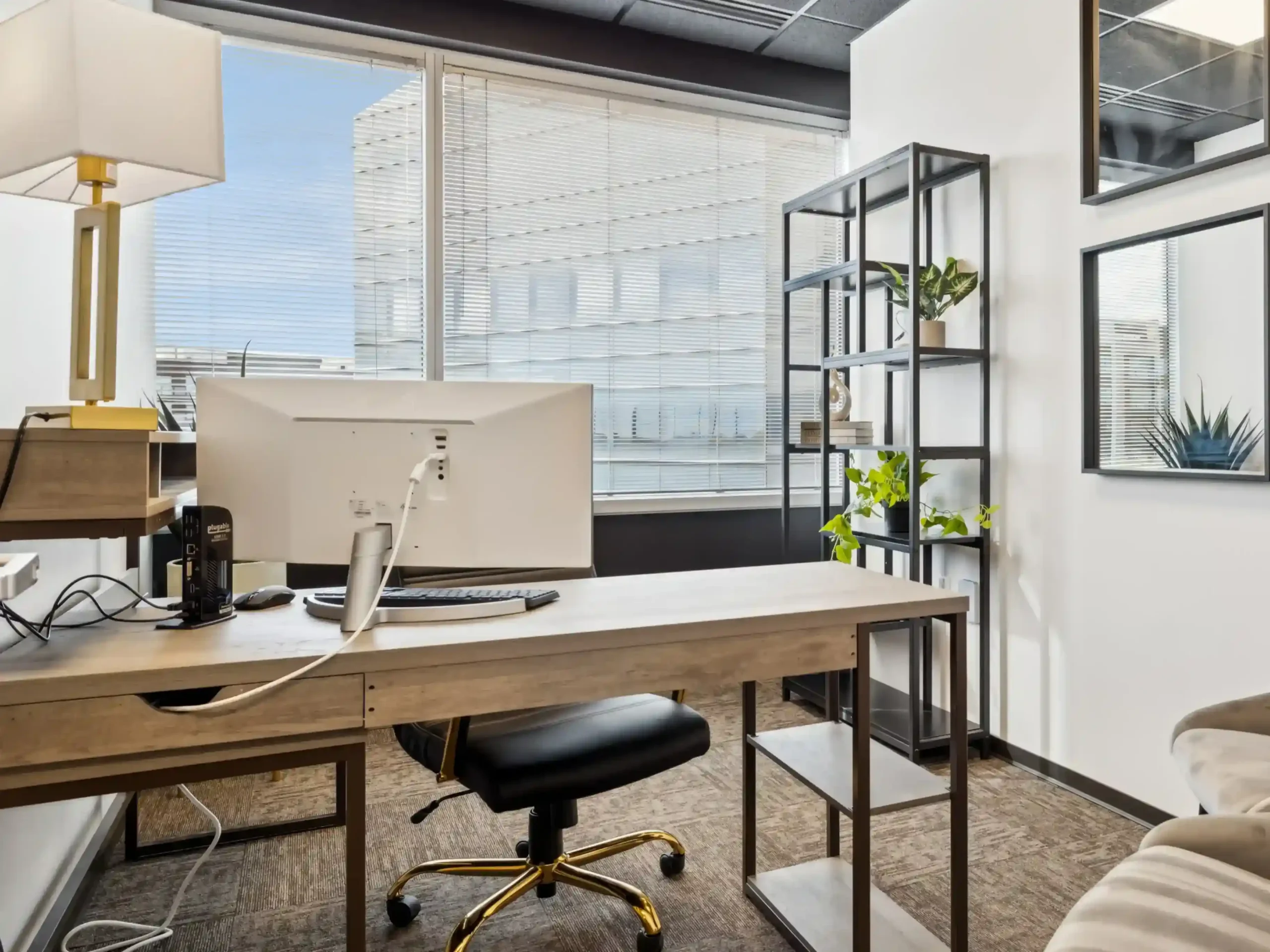 Corner office with a desk and monitor beside large windows and a black open shelving unit displaying plants and decor