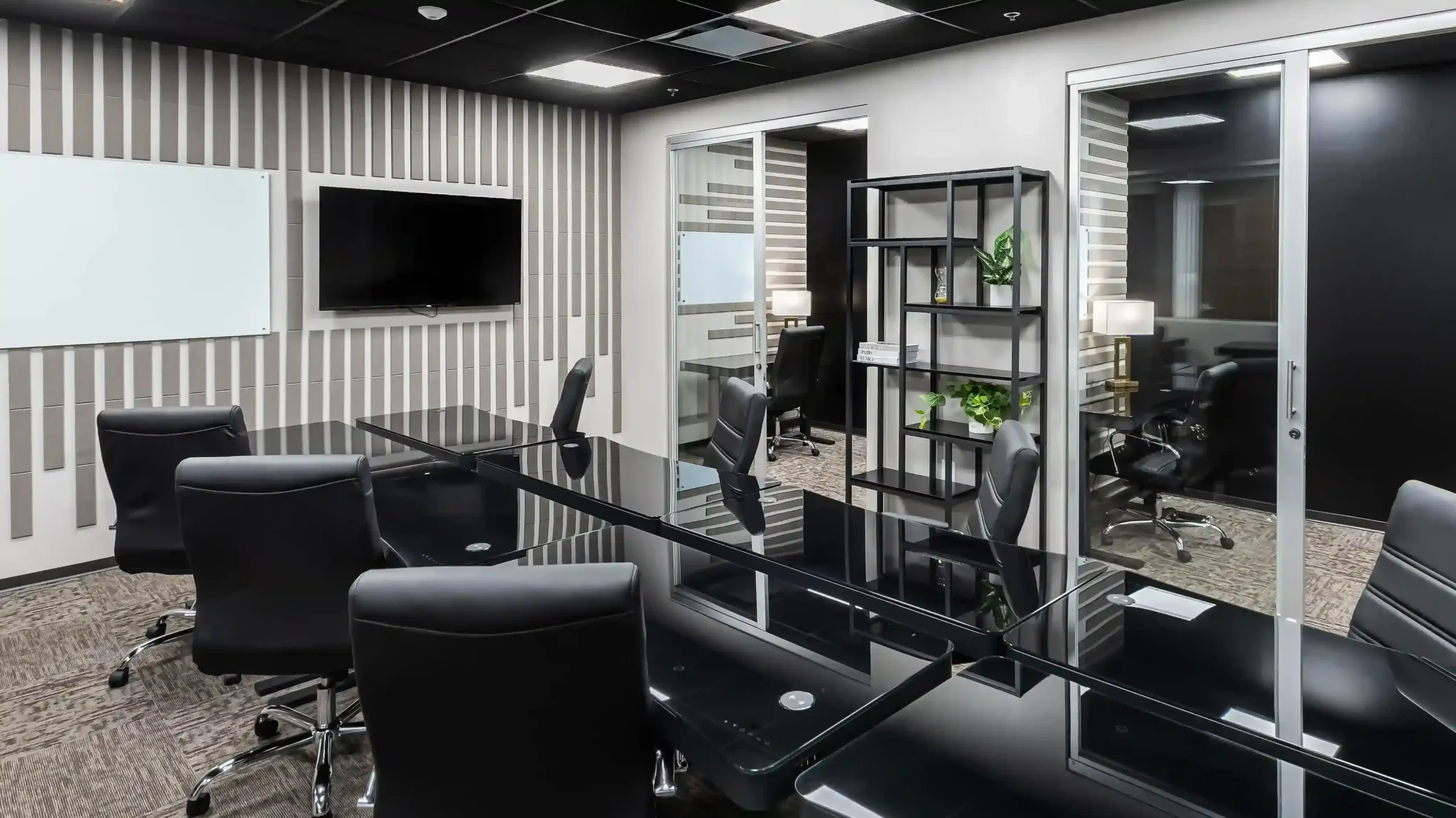 Conference room with a black table, a dark accent wall with a wall-mounted TV, and a black shelving unit holding plants