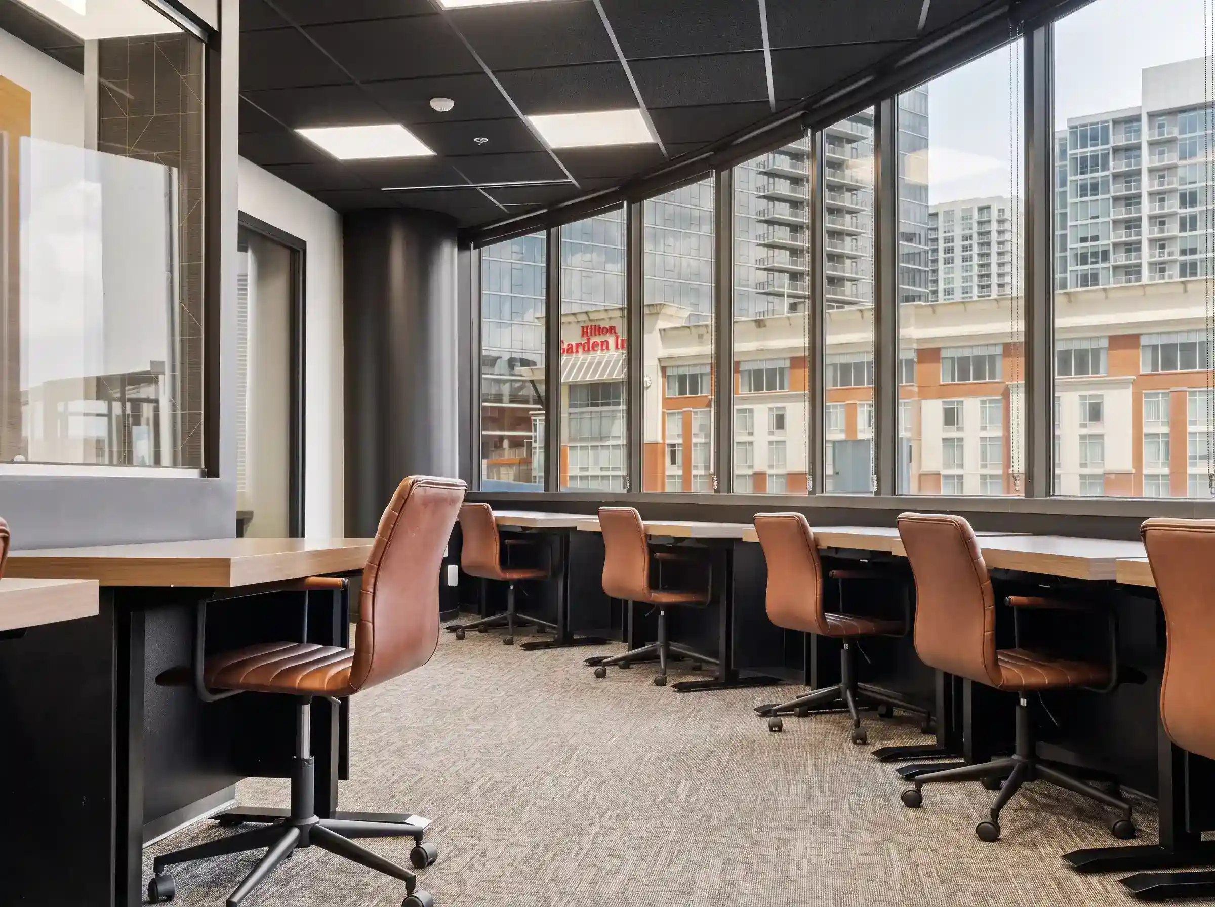 Private team office with a row of desks and brown leather task chairs along floor-to-ceiling windows overlooking city buildings